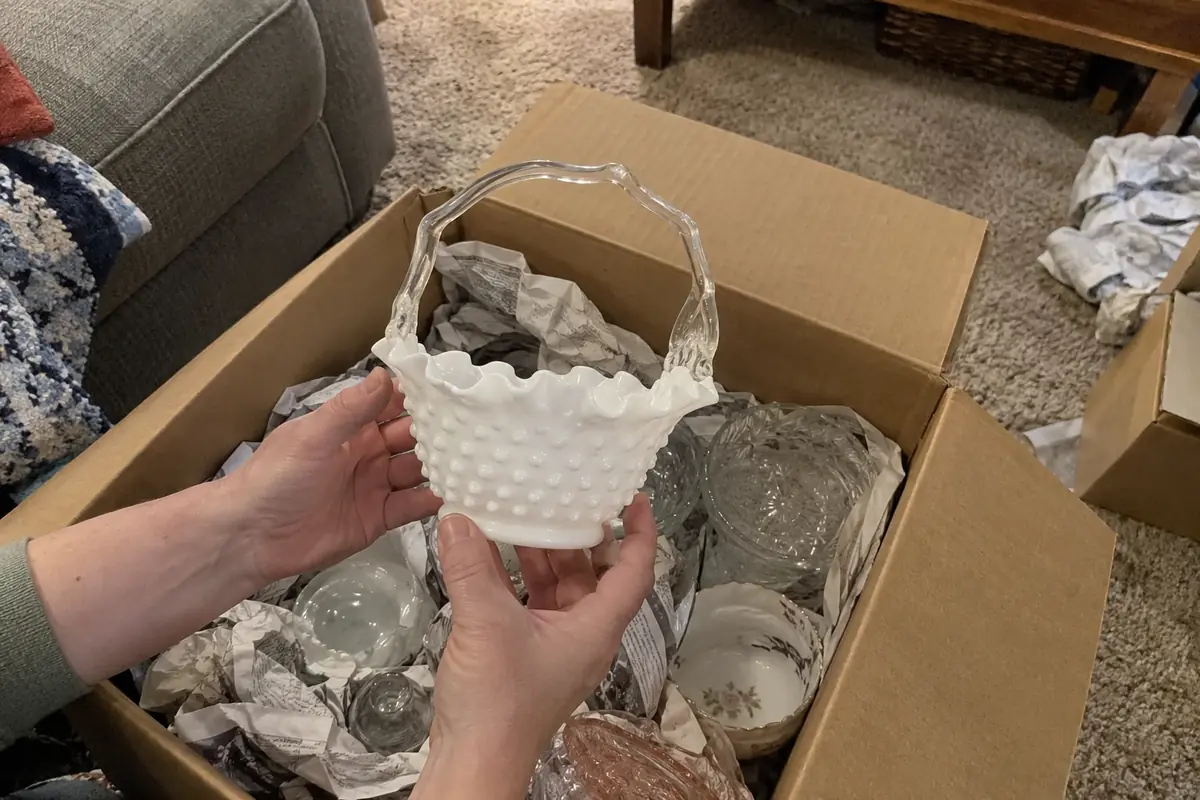 A white hobnail milk-glass basket held above a box of wrapped glassware