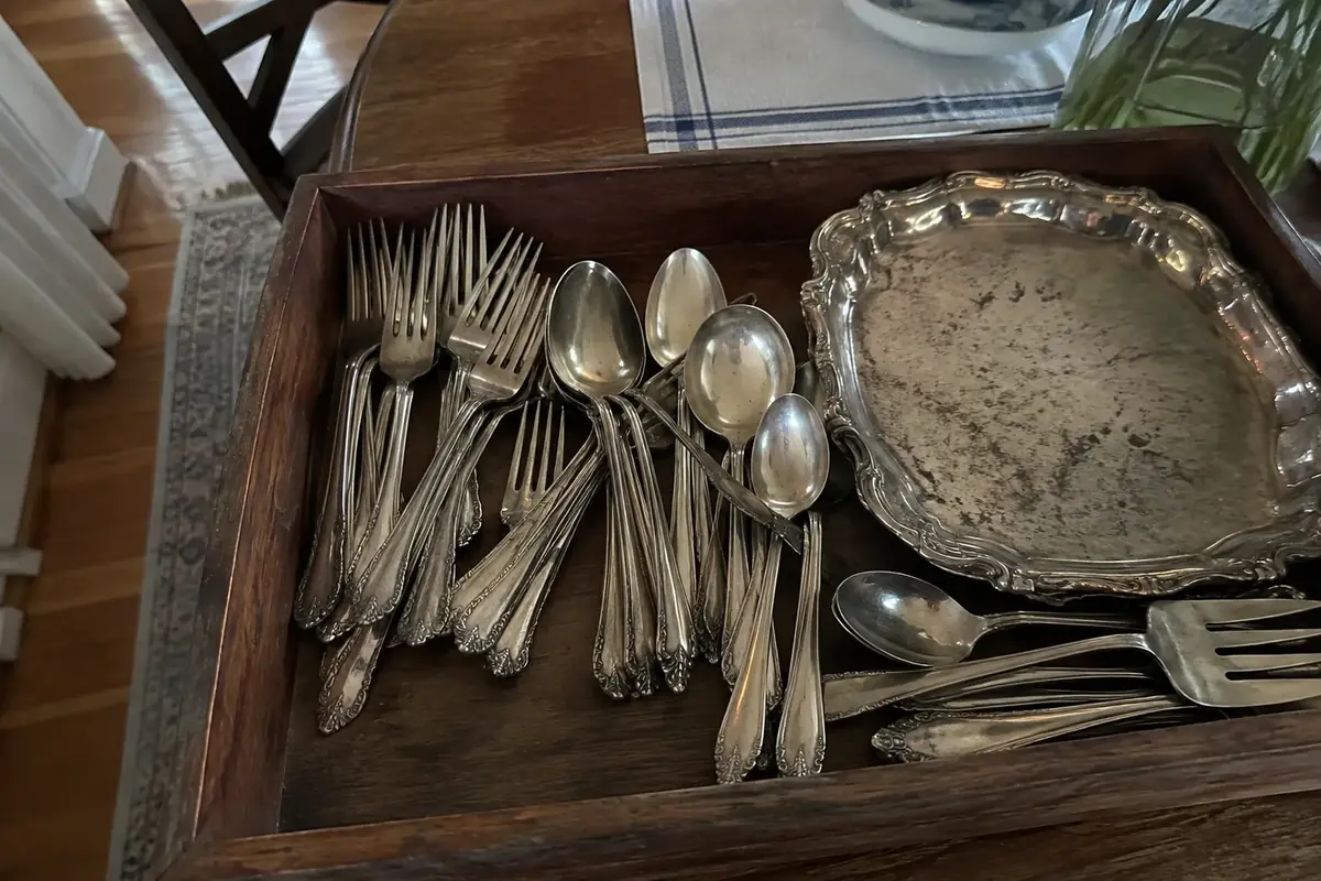 Mixed silver-plated forks, spoons and a small tray laid out on a kitchen table in daylight
